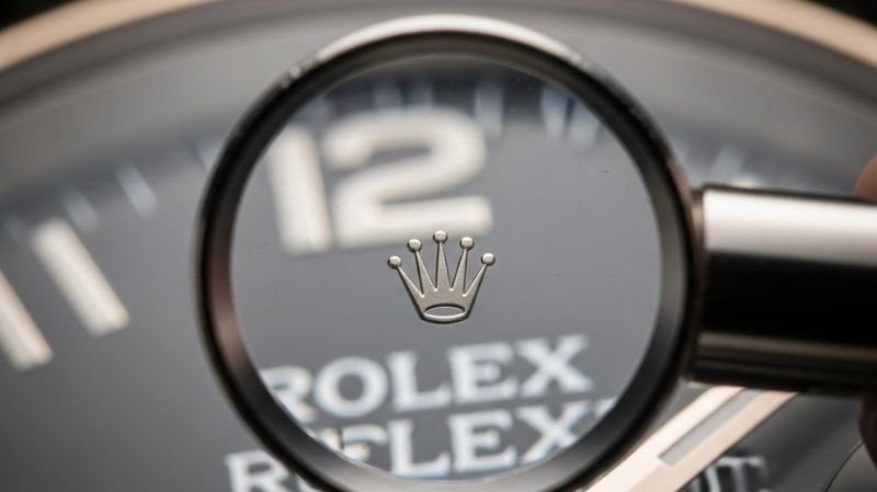 Magnified Rolex crown logo on luxury watch dial viewed through magnifying glass showing brand authentication detail.