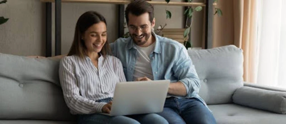 Couple sitting together on a sofa, smiling while using a laptop at home during a pre-marriage relationship course.